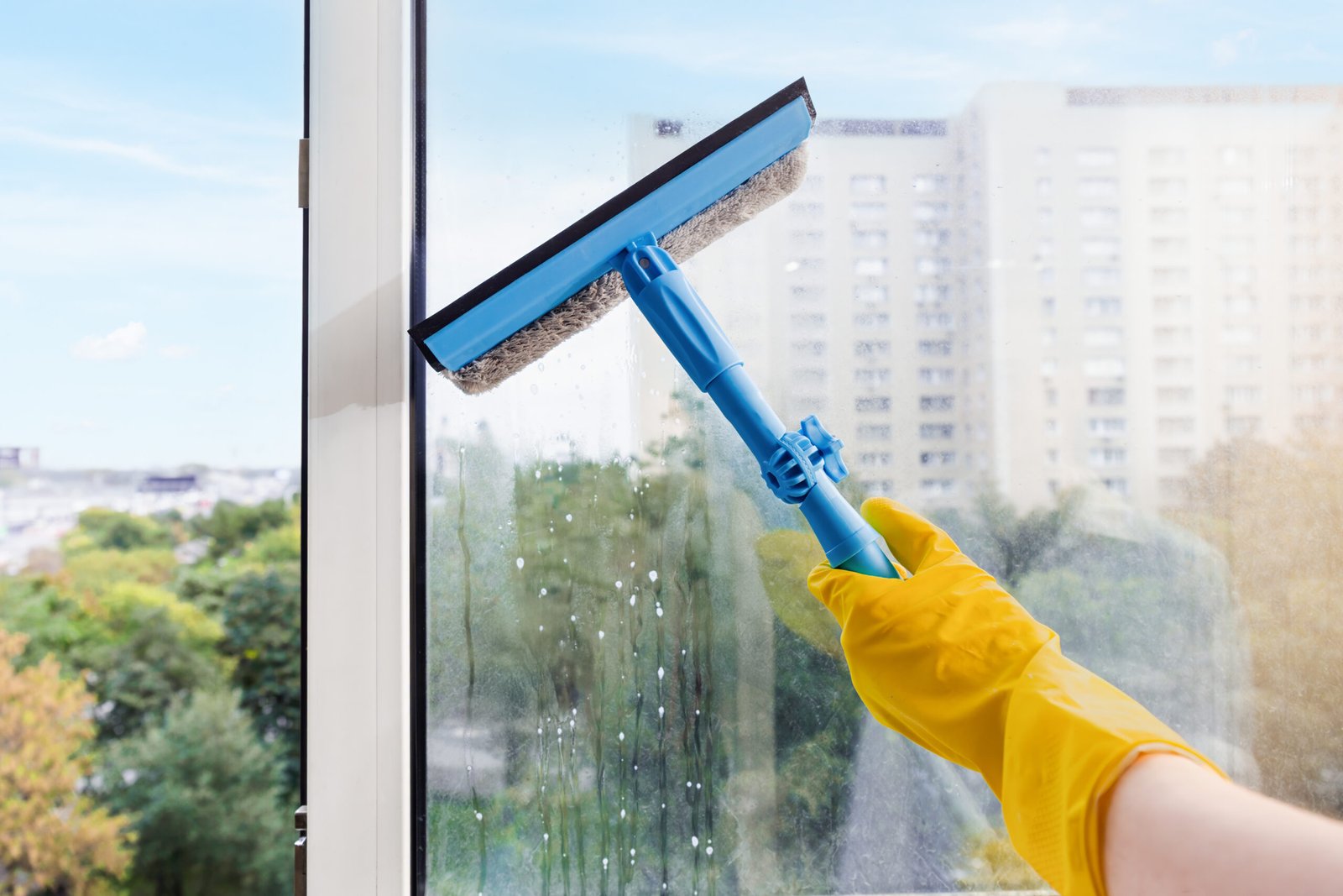 A person wearing a yellow glove uses a blue squeegee to clean a glass window, removing dirt in spring cleaning